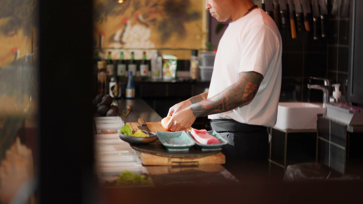 En kok i hvid T-shirt står ved sin station bag disken hos Sushi Taro