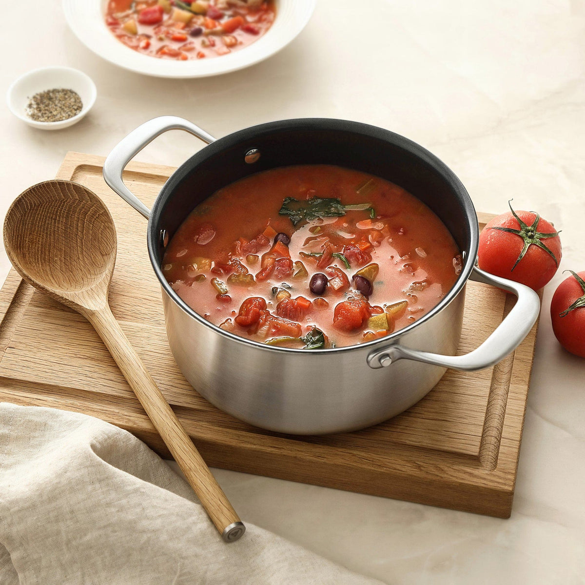 Minestrone i en gryde af rustfrit stål på et skærebræt i eg med en grydeske i træ og to tomater