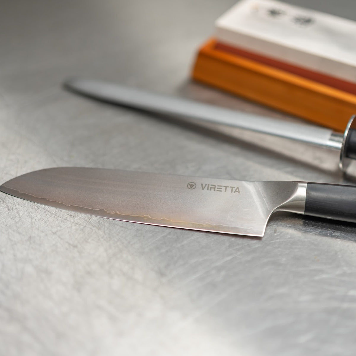 Shōjin Aira santokukniv foran et strygestål og en slibesten i bambusholder på et stålbord