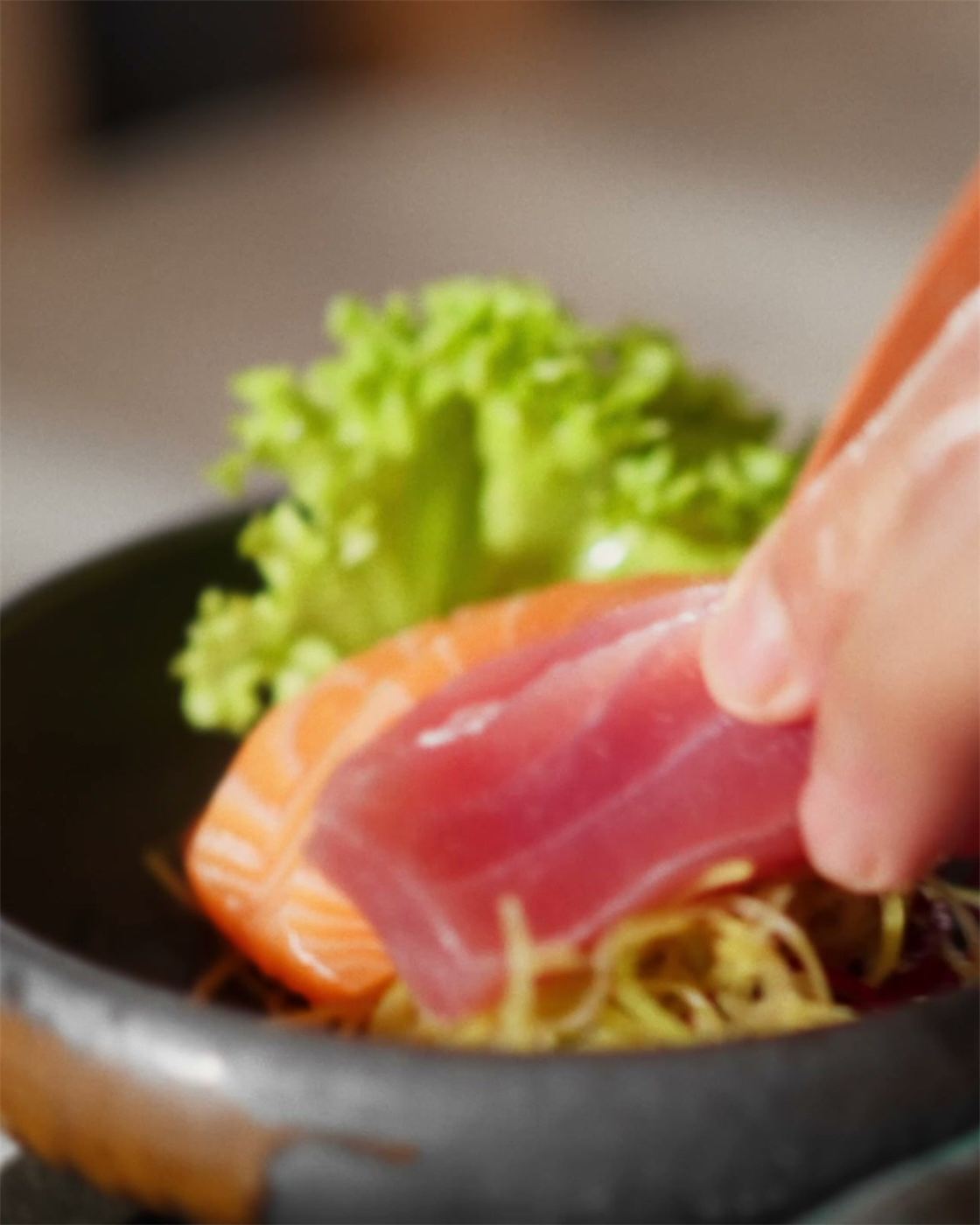 En hånd lægger et stykke tun på en mørk tallerken med laks og salat