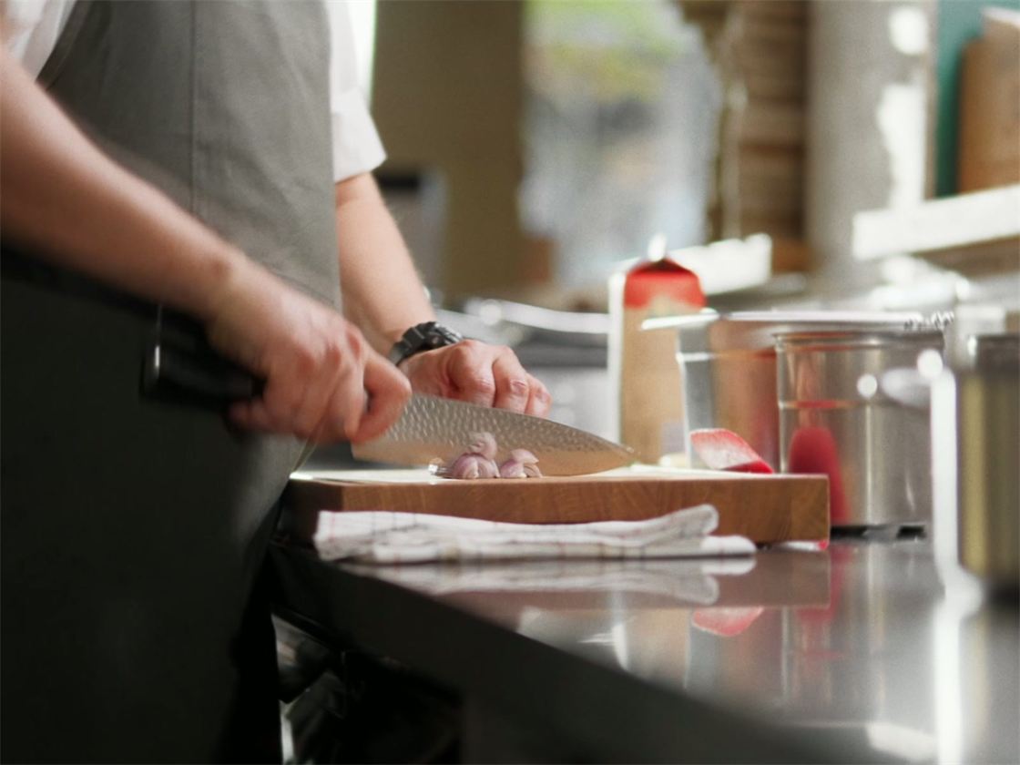 En kok i gråt forklæde hakker skalotteløg med en kniv med hamret klinge på et skærebræt i et restaurantkøkken