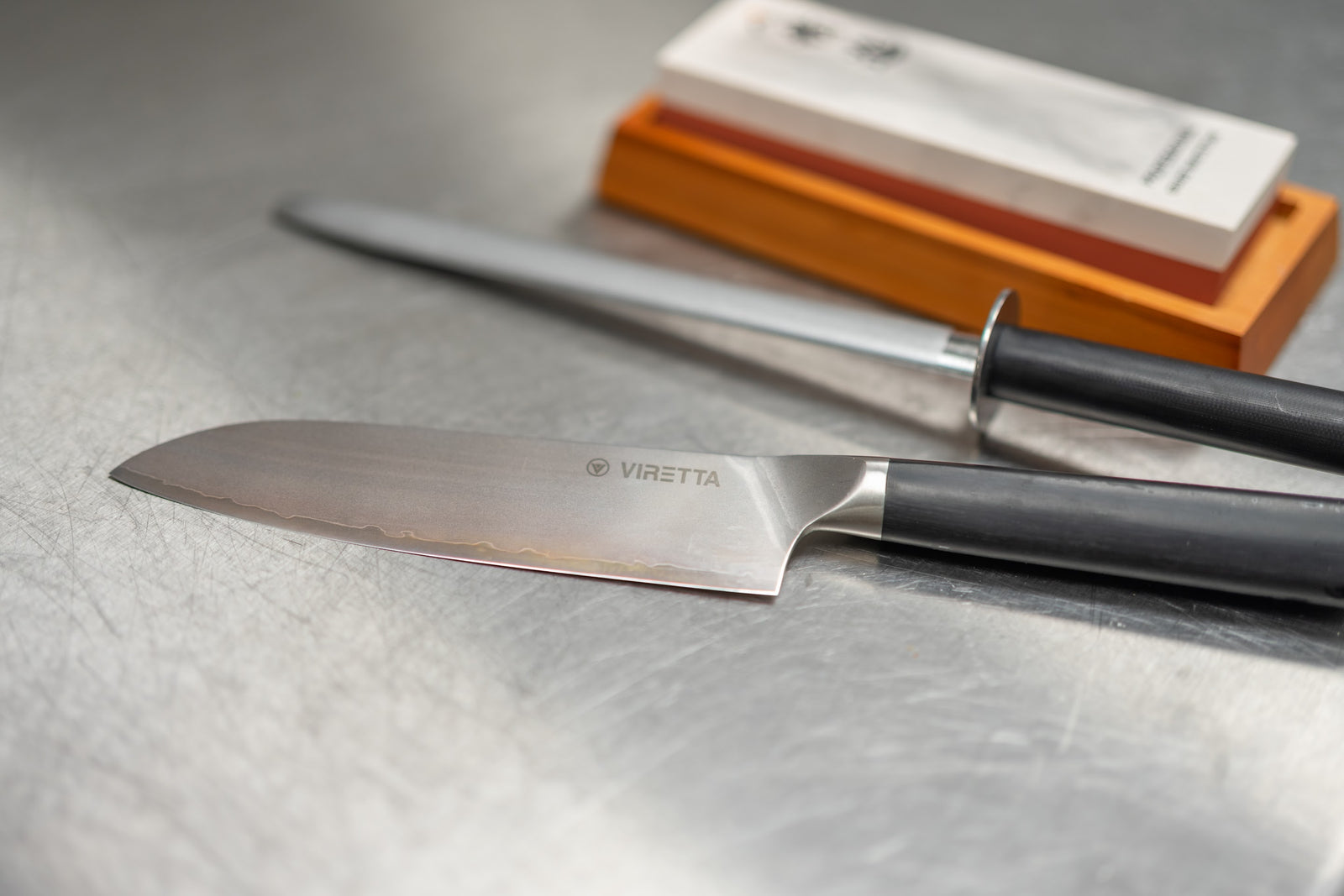 En santoku, et strygestål og en slibesten på et stålbord i køkkenet på Bolo